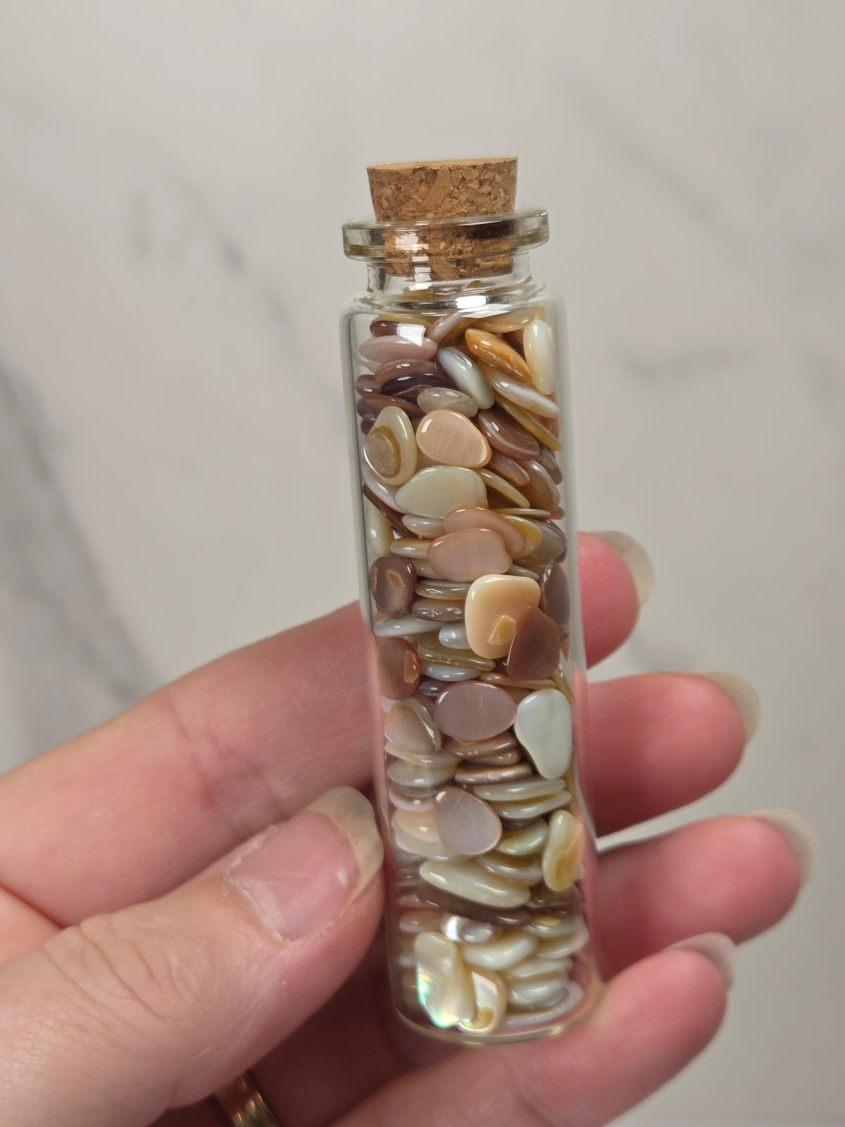 A hand holding a small clear glass vial with a cork stopper filled with smooth, polished stone chips in neutral shades of beige, cream, brown, and soft pink, against a light marble background.