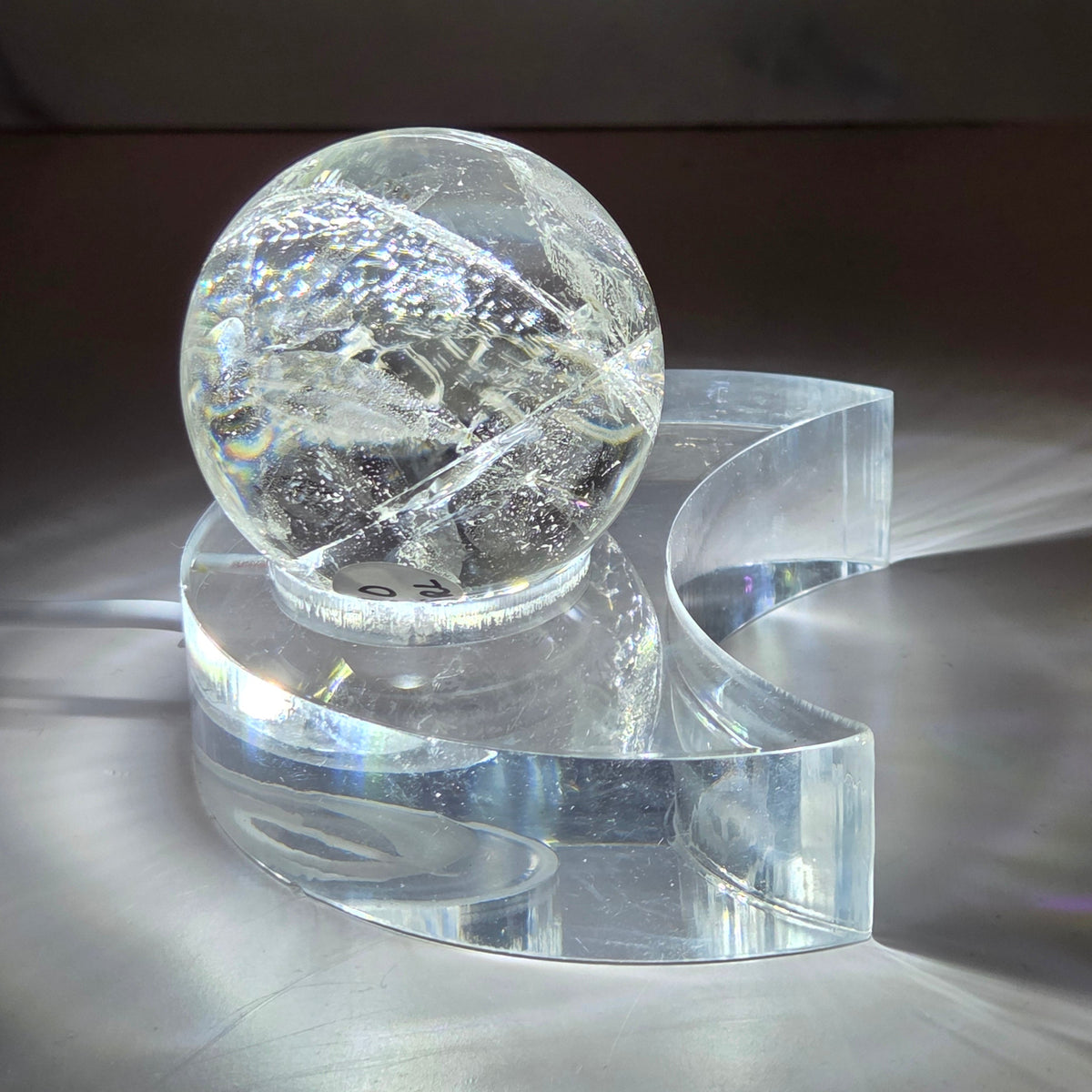 A glowing clear quartz sphere resting on a clear acrylic moon-shaped base, photographed on a white surface with light reflections around it.