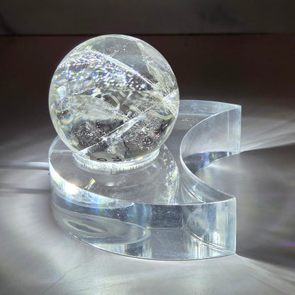A glowing clear quartz sphere resting on a clear acrylic moon-shaped base, photographed on a white surface with light reflections around it.