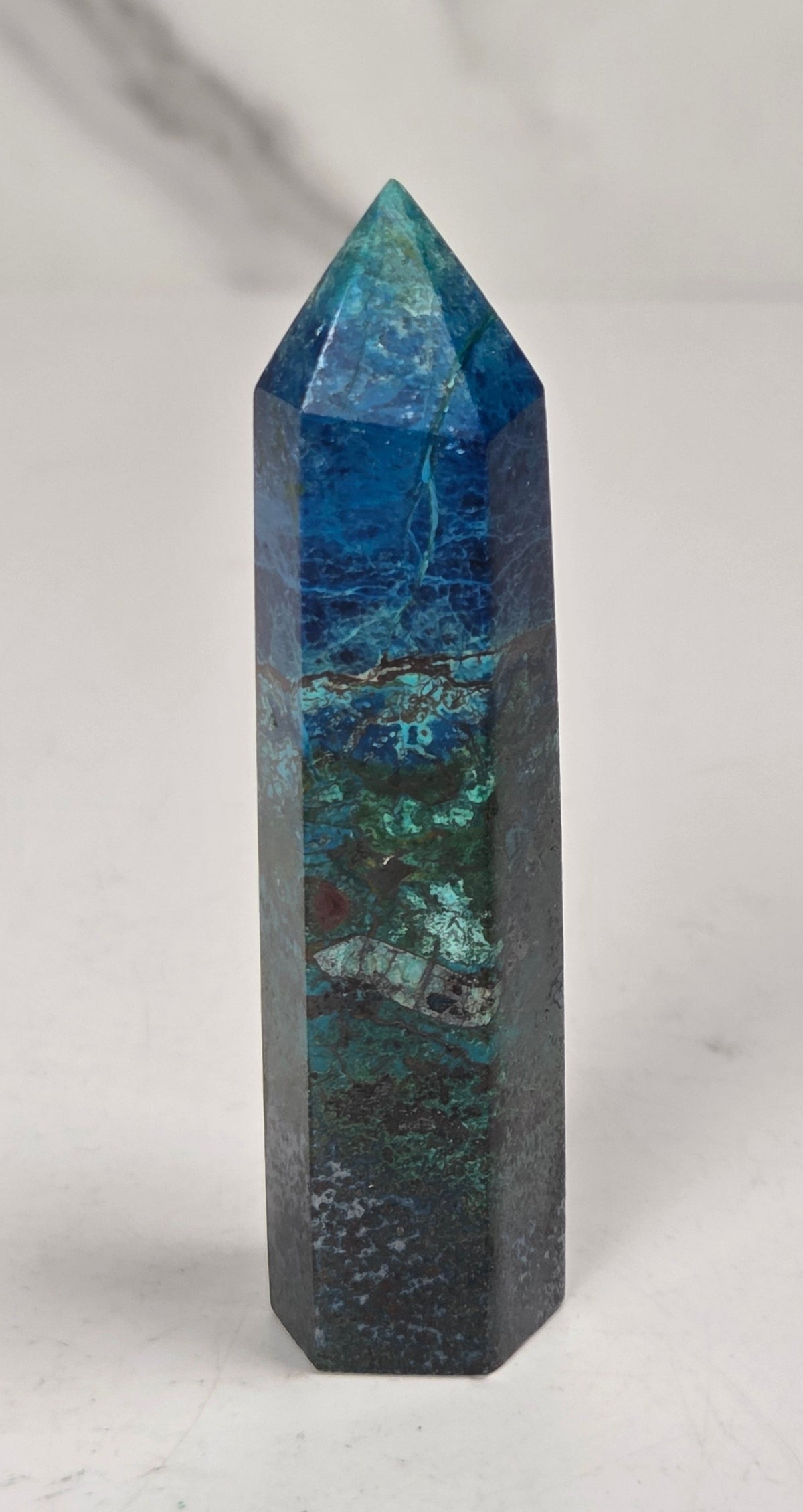 A natural blue and green jasper crystal tower point with a pointed top, standing upright on a light marble surface against a white marble background.