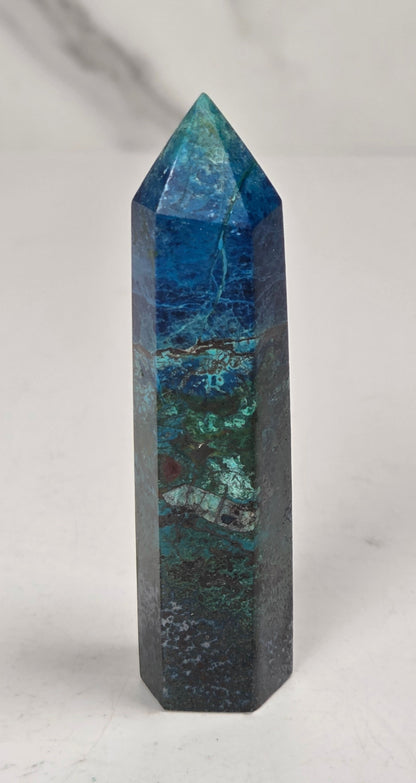A natural blue and green jasper crystal tower point with a pointed top, standing upright on a light marble surface against a white marble background.