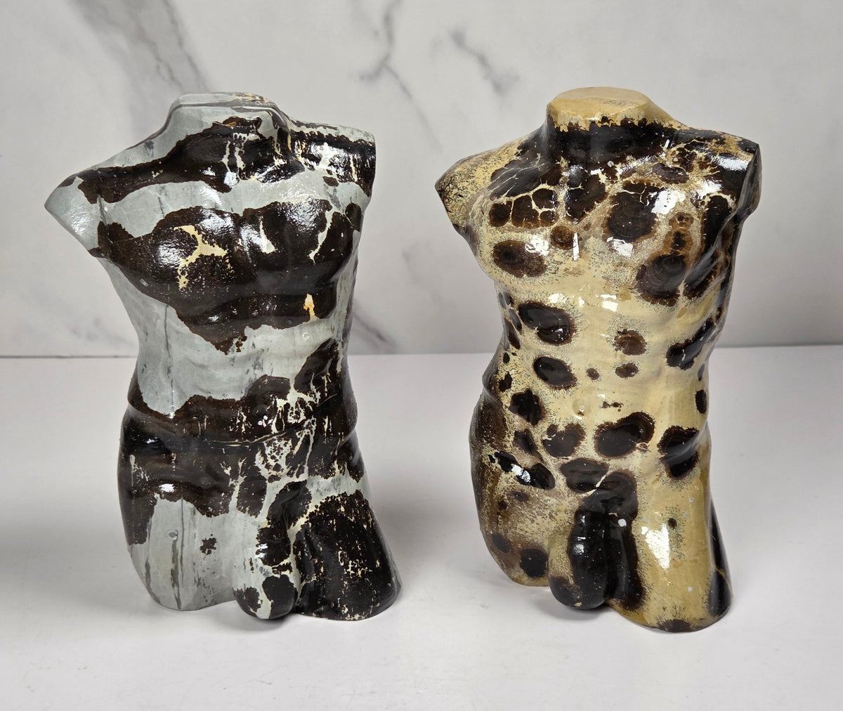 A polished stone torso sculpture with a glossy finish, featuring natural marbled patterns in reddish-brown, gray, and olive green, displayed on a white surface against a light gray marble-patterned background.