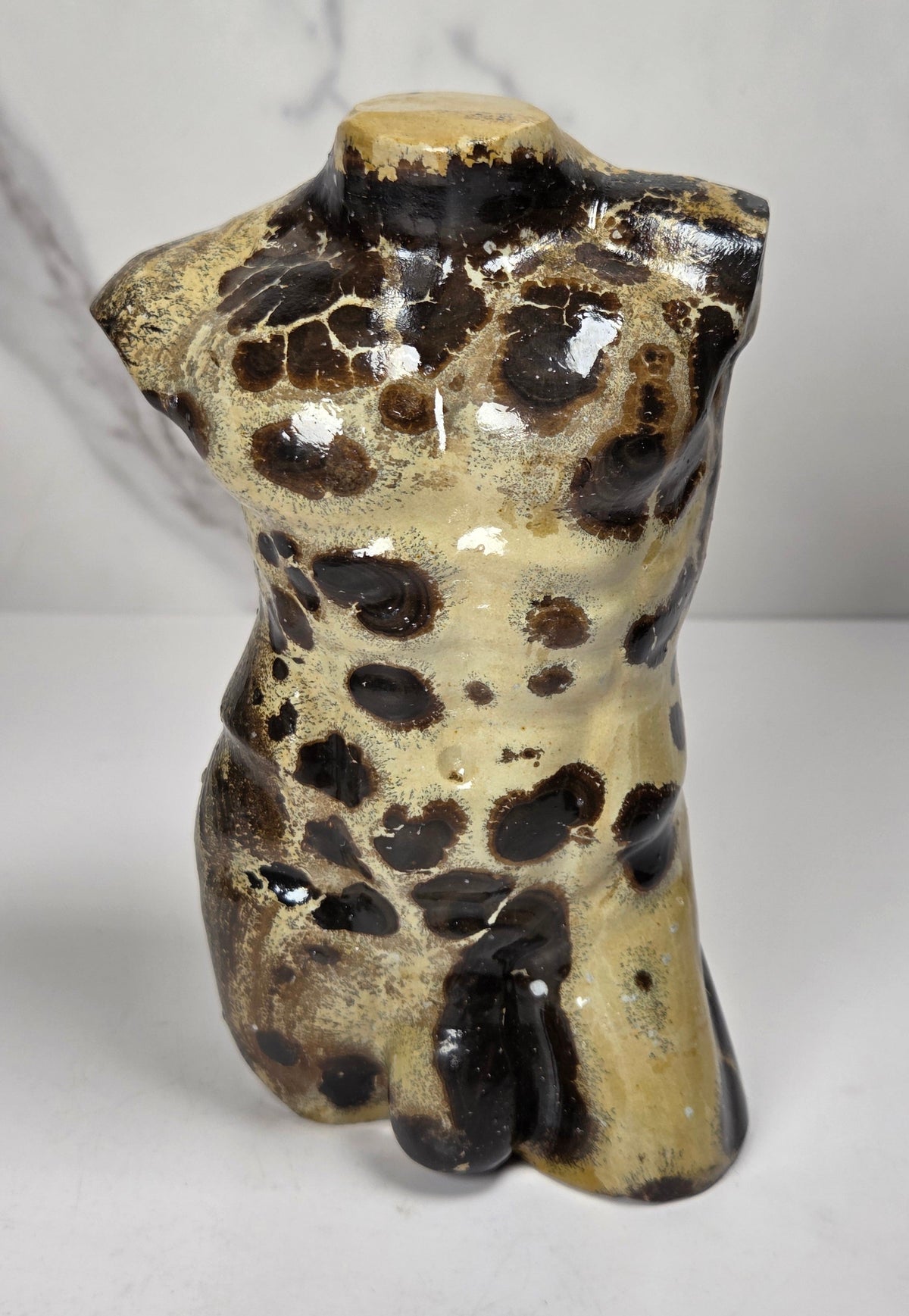 A polished stone torso sculpture with a glossy finish, featuring natural marbled patterns in reddish-brown, gray, and olive green, displayed on a white surface against a light gray marble-patterned background.