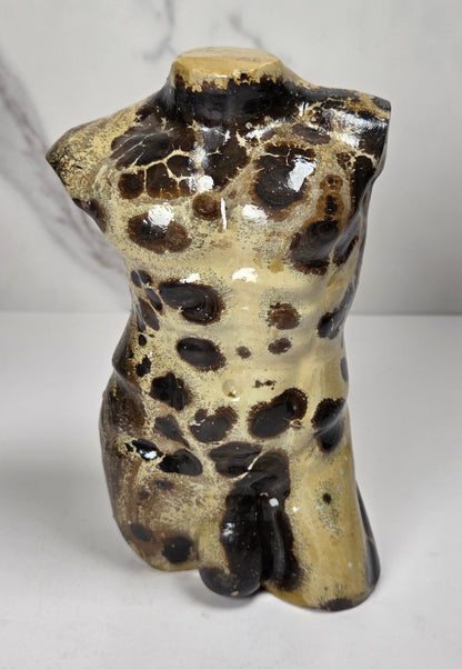 A polished stone torso sculpture with a glossy finish, featuring natural marbled patterns in reddish-brown, gray, and olive green, displayed on a white surface against a light gray marble-patterned background.