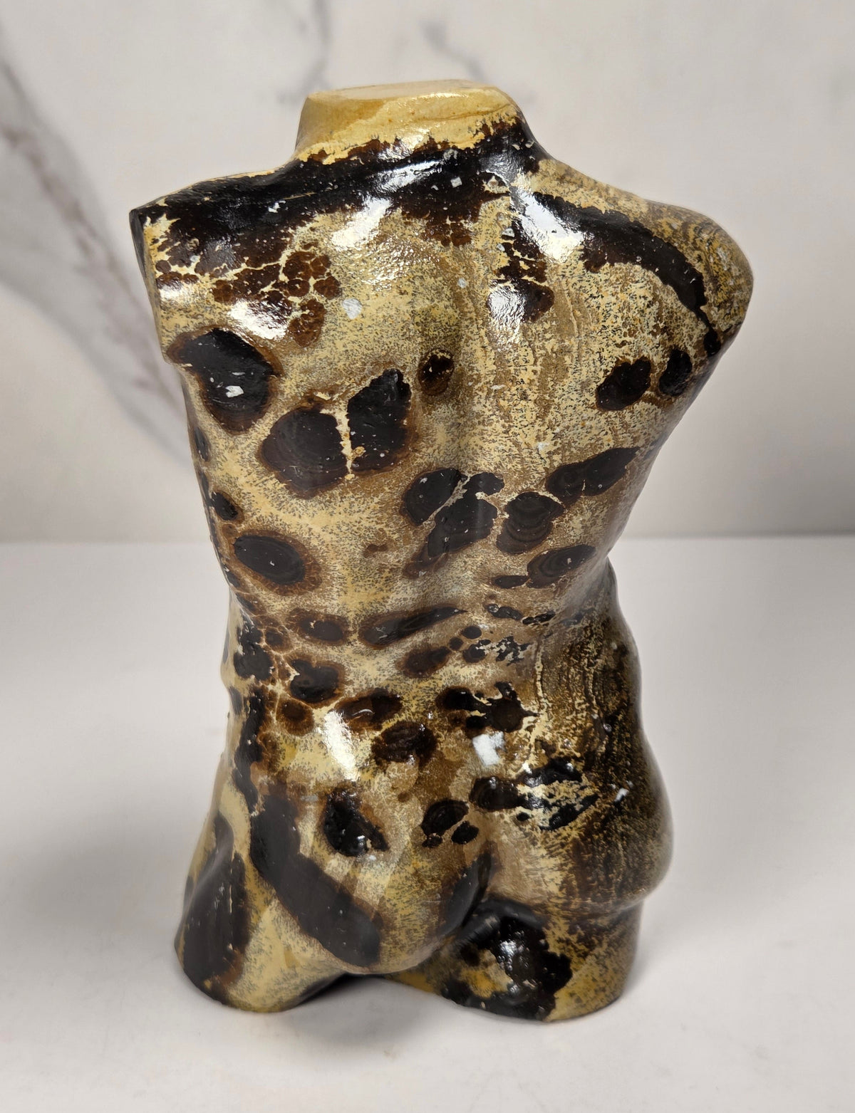 A polished stone torso sculpture with a glossy finish, featuring natural marbled patterns in reddish-brown, gray, and olive green, displayed on a white surface against a light gray marble-patterned background.