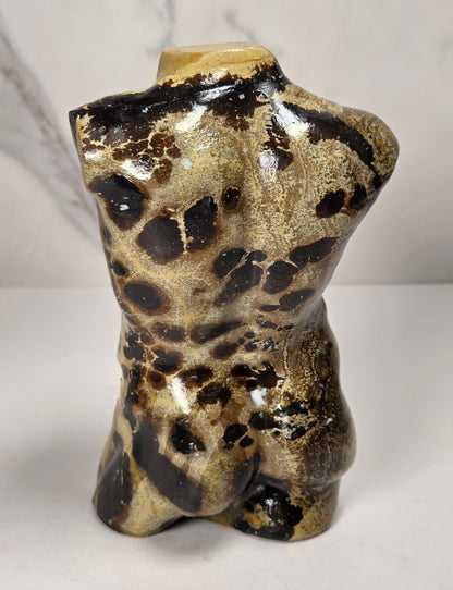 A polished stone torso sculpture with a glossy finish, featuring natural marbled patterns in reddish-brown, gray, and olive green, displayed on a white surface against a light gray marble-patterned background.