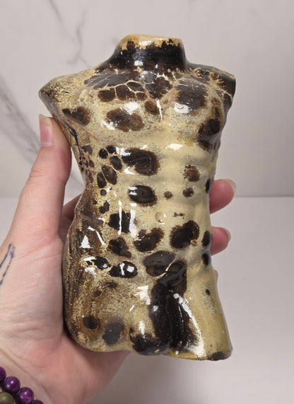 Hand holding a polished stone torso sculpture with a glossy finish, featuring natural marbled patterns in reddish-brown, gray, and olive green, displayed on a white surface against a light gray marble-patterned background.