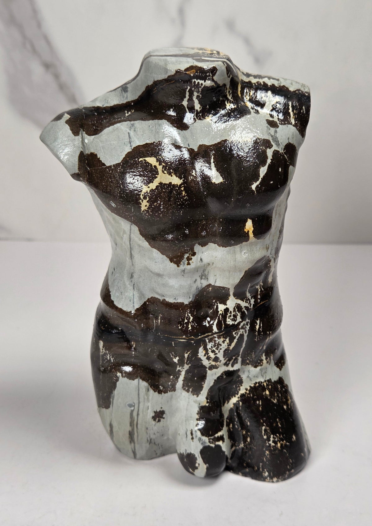 A polished stone torso sculpture with a glossy finish, featuring natural marbled patterns in reddish-brown, gray, and olive green, displayed on a white surface against a light gray marble-patterned background.