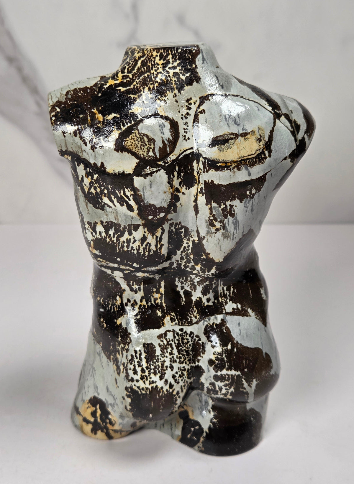 A polished stone torso sculpture with a glossy finish, featuring natural marbled patterns in reddish-brown, gray, and olive green, displayed on a white surface against a light gray marble-patterned background.
