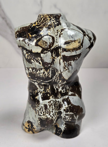 A polished stone torso sculpture with a glossy finish, featuring natural marbled patterns in reddish-brown, gray, and olive green, displayed on a white surface against a light gray marble-patterned background.