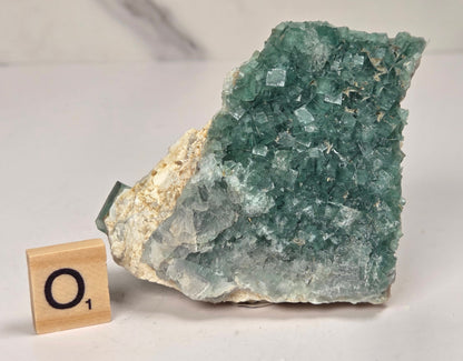 Green Fluorite specimen with a wood tile with the letter O against a marbled background