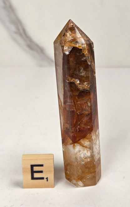 A natural clear quartz point crystal with prominent golden-brown and amber inclusions, displayed next to a wooden '
E1' tile on a light gray marble surface.