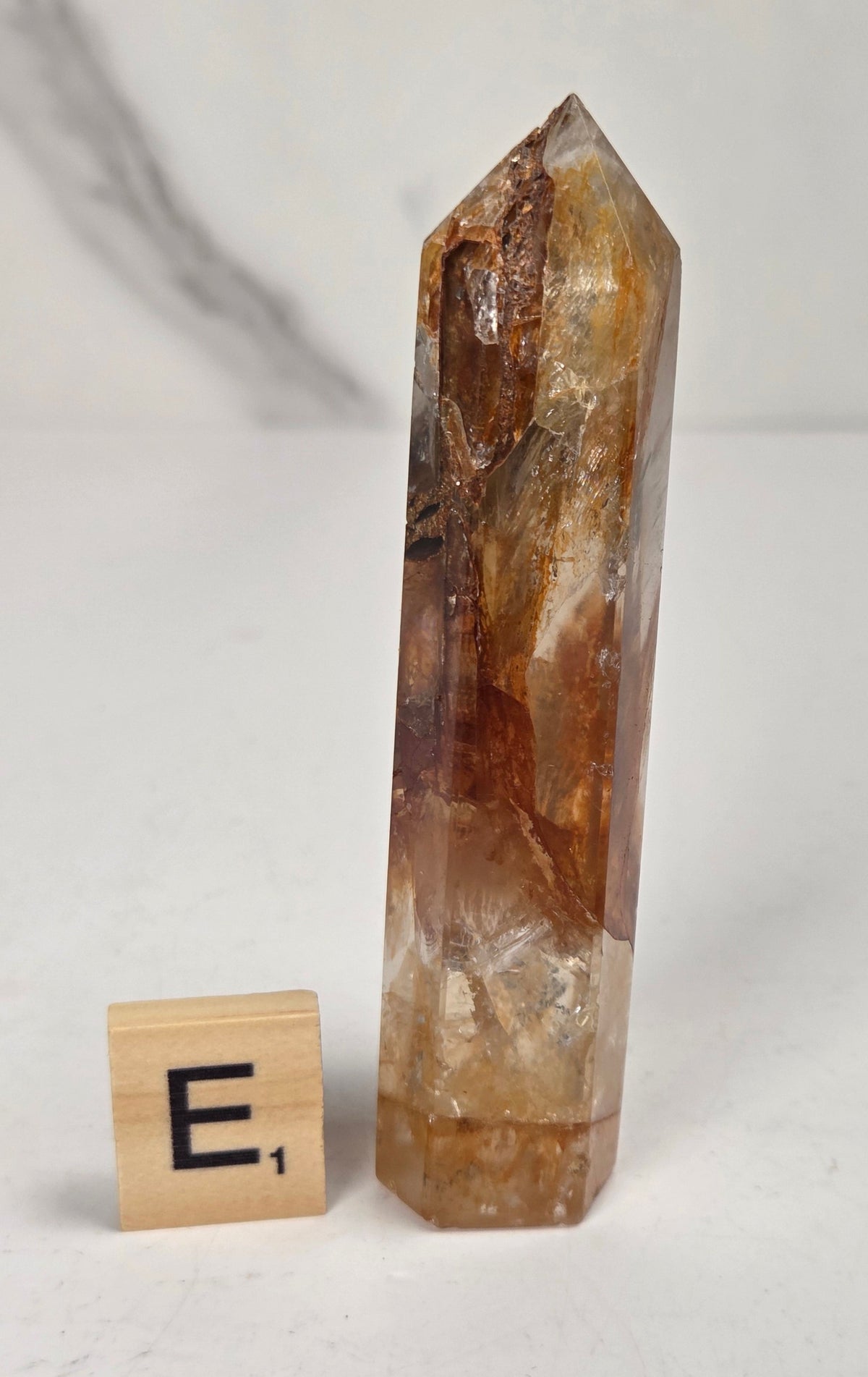 A natural clear quartz point crystal with prominent golden-brown and amber inclusions, displayed next to a wooden '
E1' tile on a light gray marble surface.