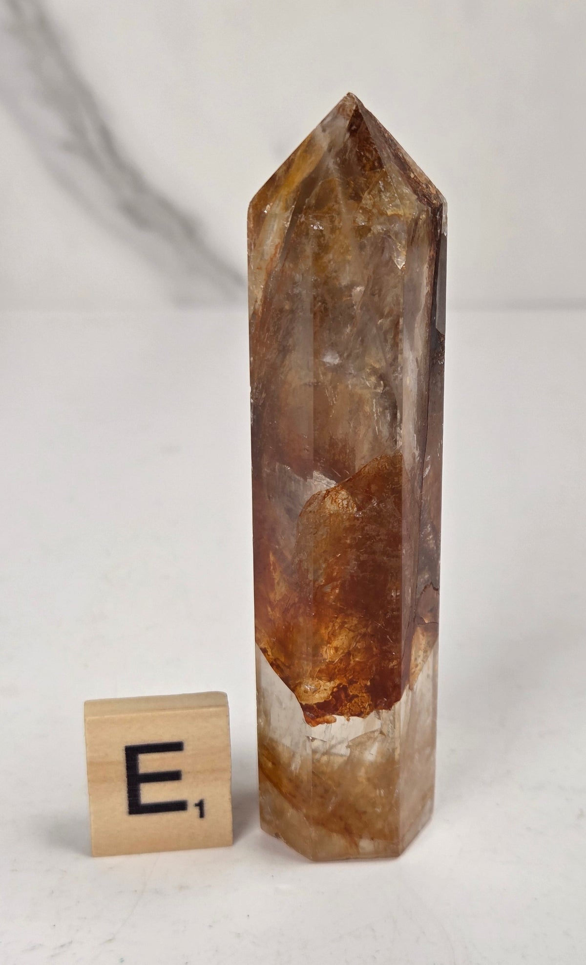 A natural clear quartz point crystal with prominent golden-brown and amber inclusions, displayed next to a wooden '
E1' tile on a light gray marble surface.