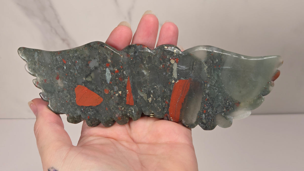 Hand holding a crystal heart with wings carving with black, grey, red and gold coloring against a white background