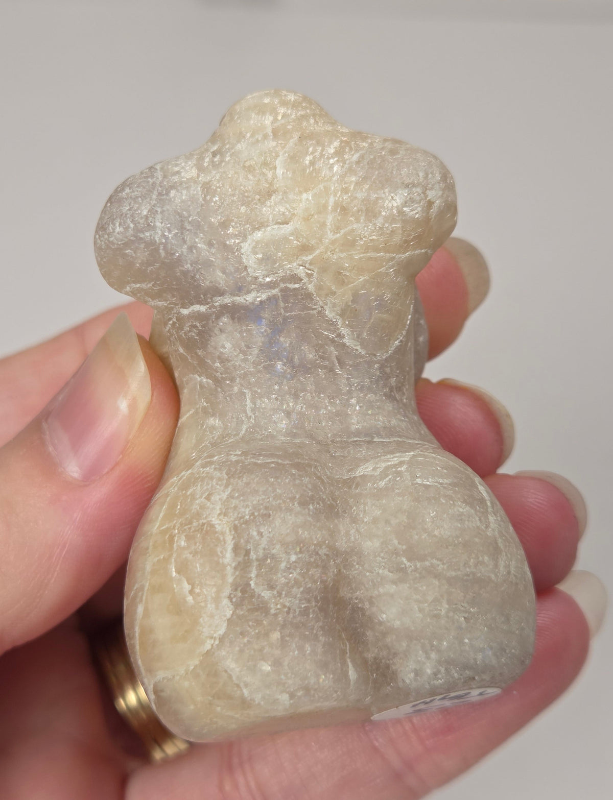 A hand holding a small carved stone figurine with a pearlescent, milky appearance and subtle blue iridescence, set against a light marble-patterned background.