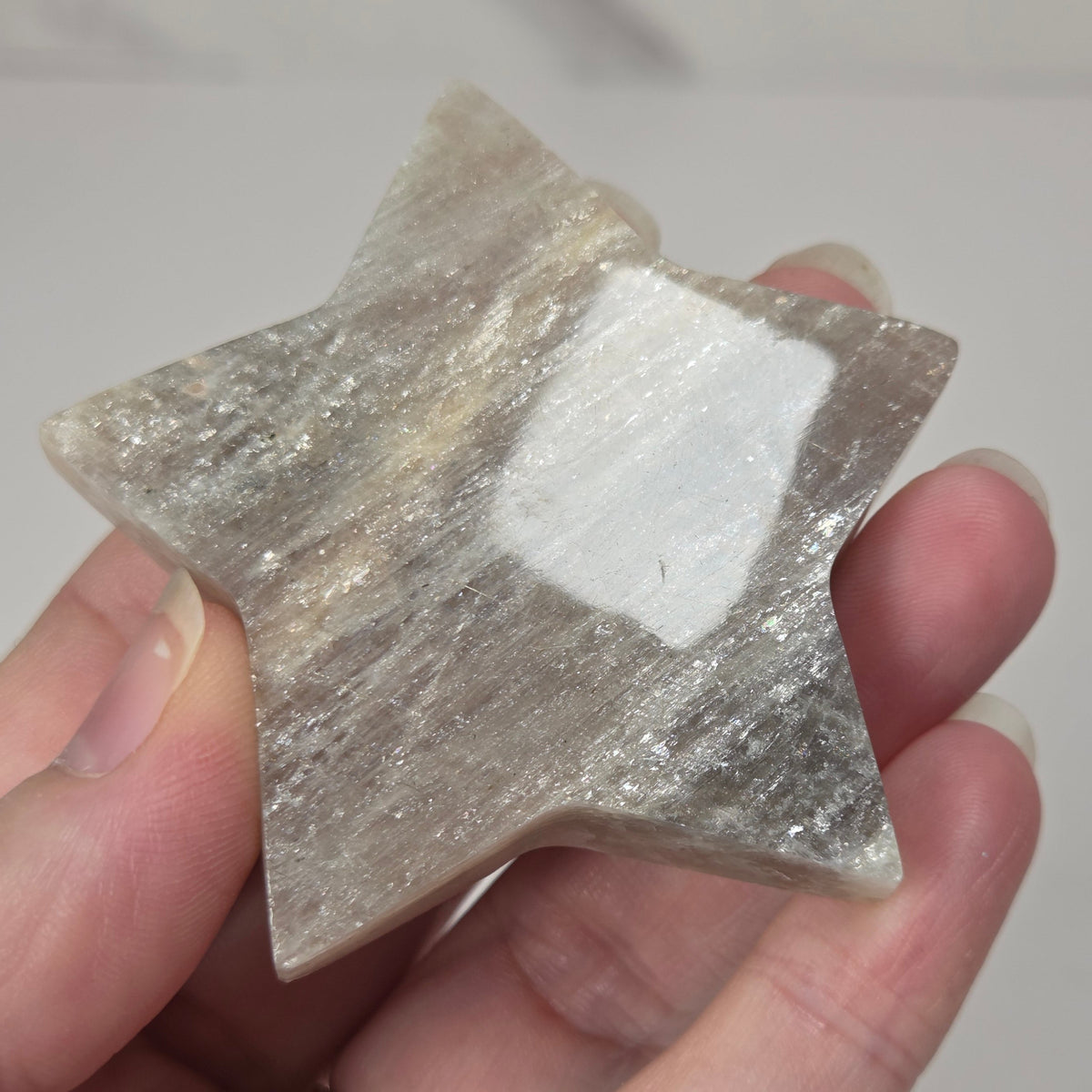 crystal star shaped carving made of moonstone showing white flash being held in a hand against a white background
