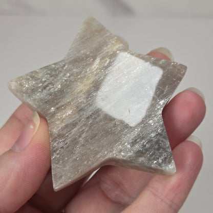crystal star shaped carving made of moonstone showing white flash being held in a hand against a white background