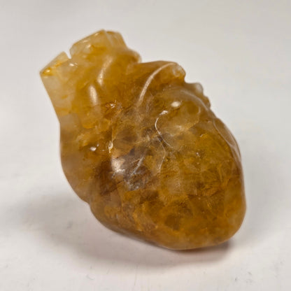A small carved translucent heart-shaped stone pendant with gold coloring, photographed against a softly blurred light background.