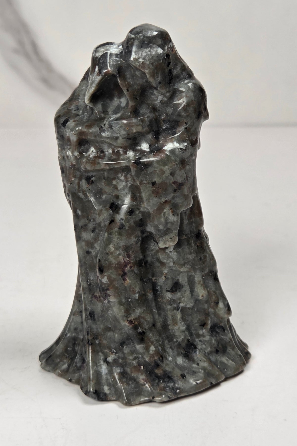 A black textured sculpture of a hooded figure, resembling a grim reaper or raven, standing on a white surface against a light gray marble-patterned background.