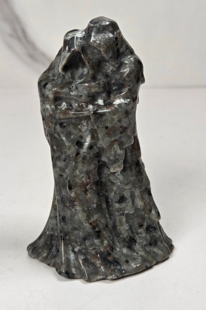A black textured sculpture of a hooded figure, resembling a grim reaper or raven, standing on a white surface against a light gray marble-patterned background.