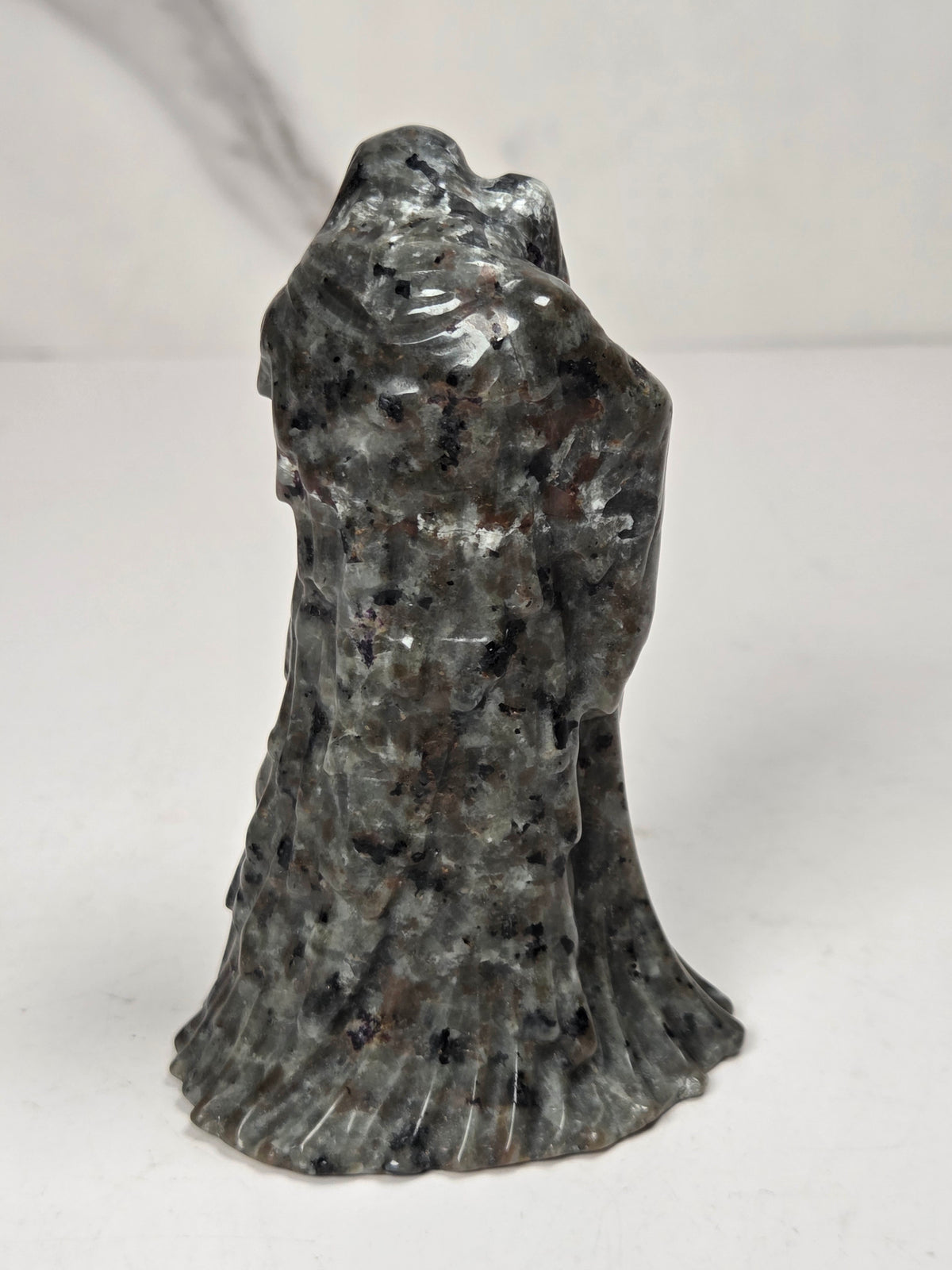 A black textured sculpture of a hooded figure, resembling a grim reaper or raven, standing on a white surface against a light gray marble-patterned background.