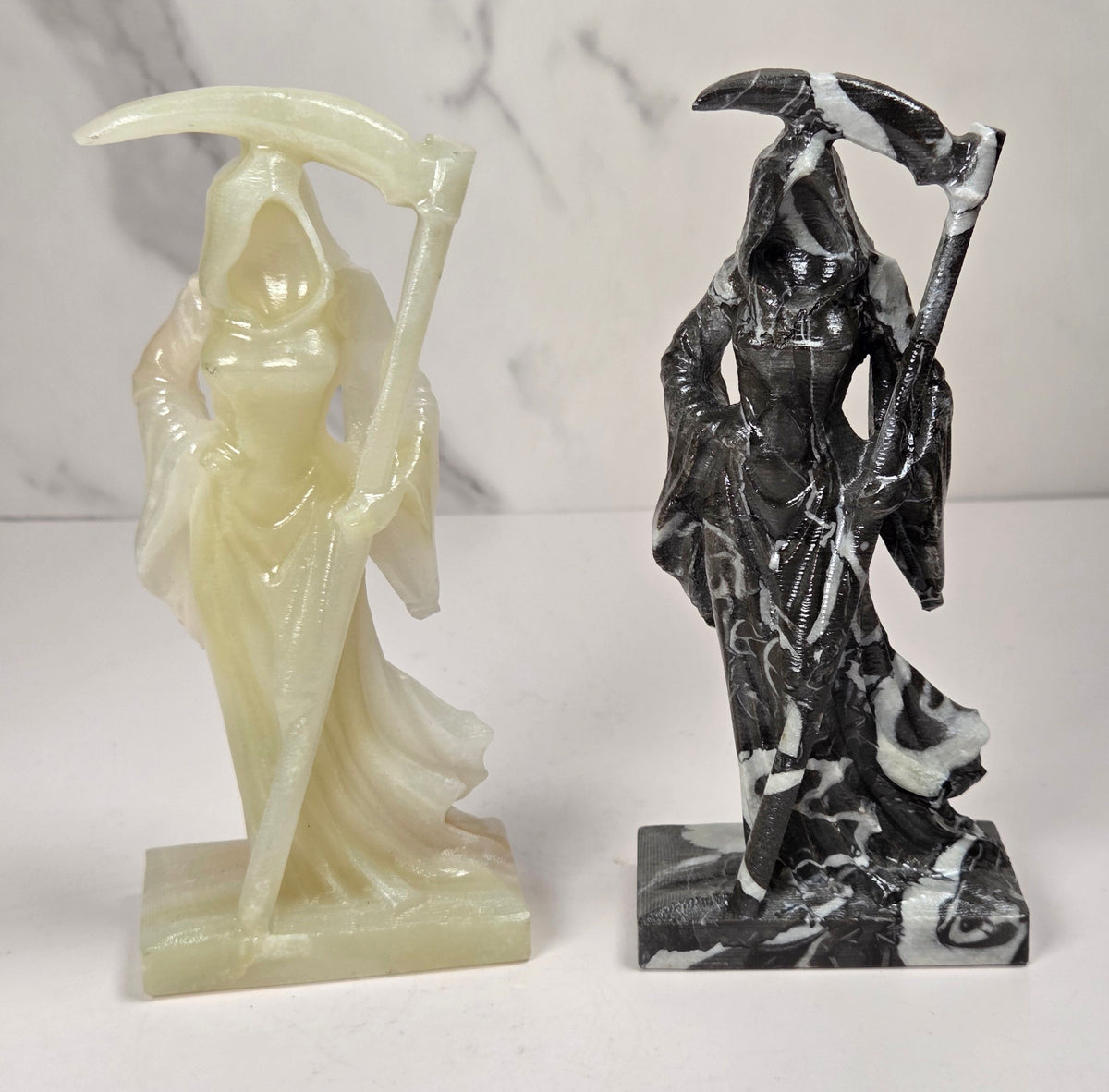 faceless lady reaper carvings made of a golden tan color and a marbled black and white mineral against a marbled white background