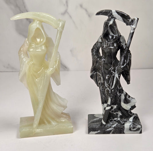 faceless lady reaper carvings made of a golden tan color and a marbled black and white mineral against a marbled white background