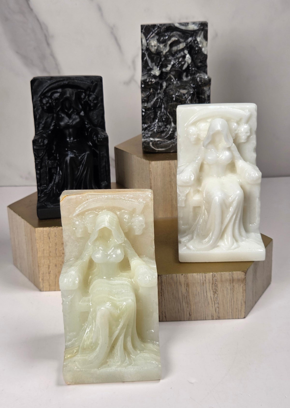 Carvings of a faceless woman wearing a robe, sitting on a chair, in various colors, on a wooden stand against a marble background