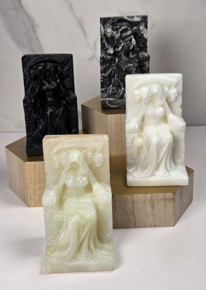 Carvings of a faceless woman wearing a robe, sitting on a chair, in various colors, on a wooden stand against a marble background