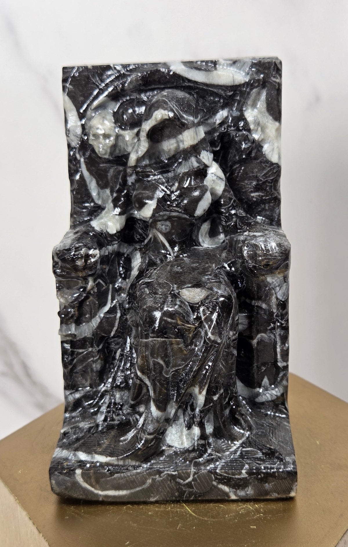 Carving of a faceless woman wearing a robe, sitting on a chair, in marbled black and white, on a wooden stand against a marble background
