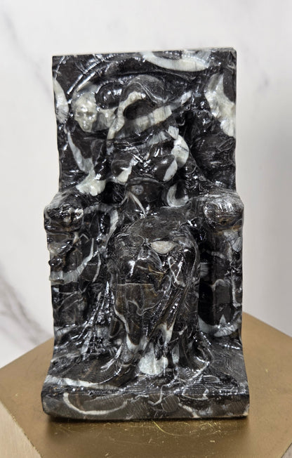 Carving of a faceless woman wearing a robe, sitting on a chair, in marbled black and white, on a wooden stand against a marble background
