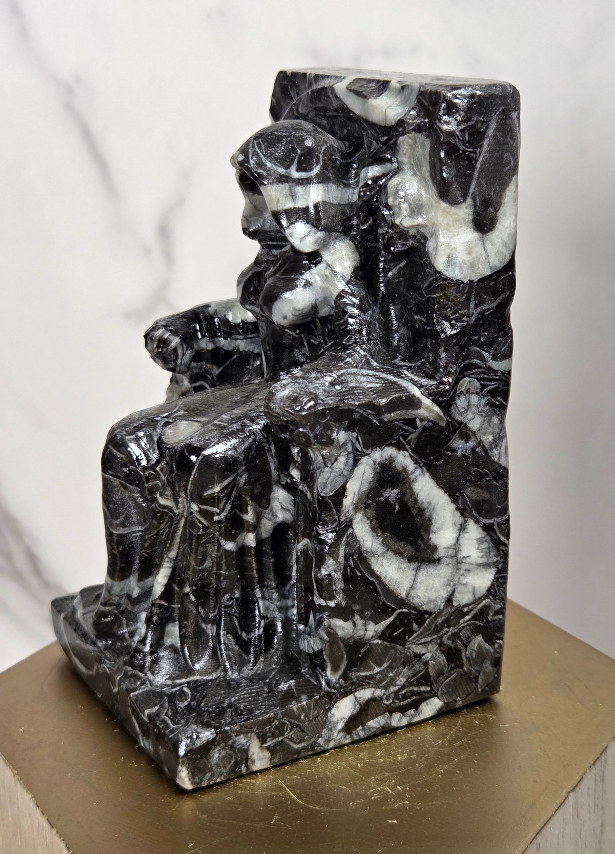 Carving of a faceless woman wearing a robe, sitting on a chair, in marbled black and white, on a wooden stand against a marble background