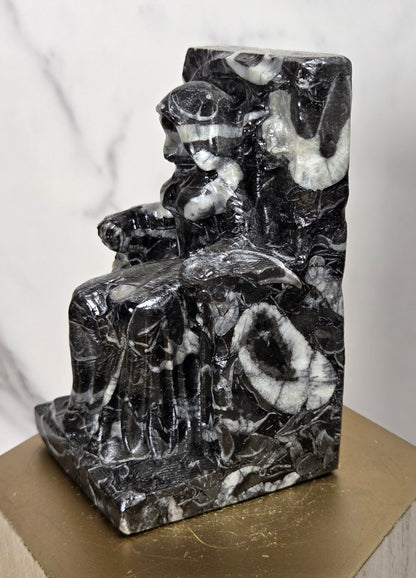 Carving of a faceless woman wearing a robe, sitting on a chair, in marbled black and white, on a wooden stand against a marble background