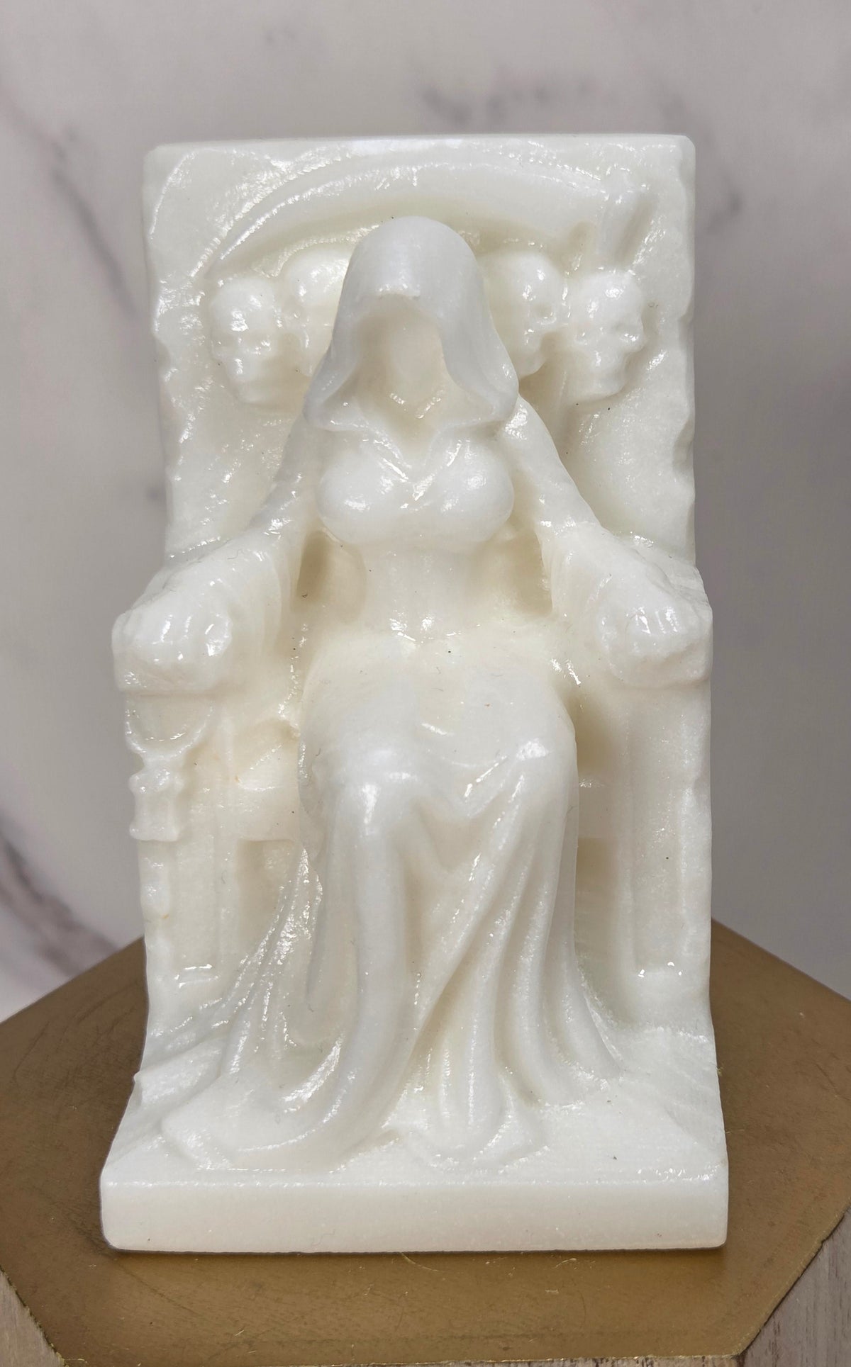 Carving of a faceless woman wearing a robe, sitting on a chair, in white jade, on a wooden stand against a marble background