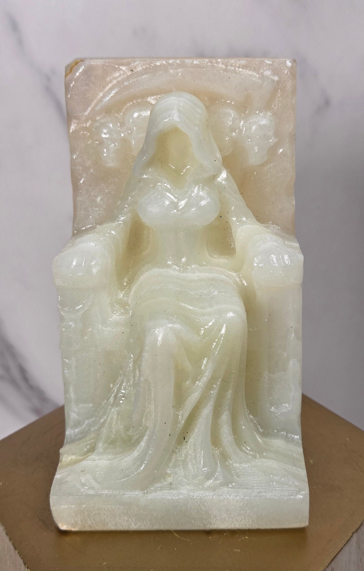 Carving of a faceless woman wearing a robe, sitting on a chair, in a striped crystal, on a wooden stand against a marble background