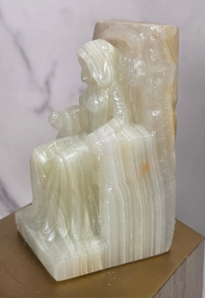 Carving of a faceless woman wearing a robe, sitting on a chair, in a striped crystal, on a wooden stand against a marble background