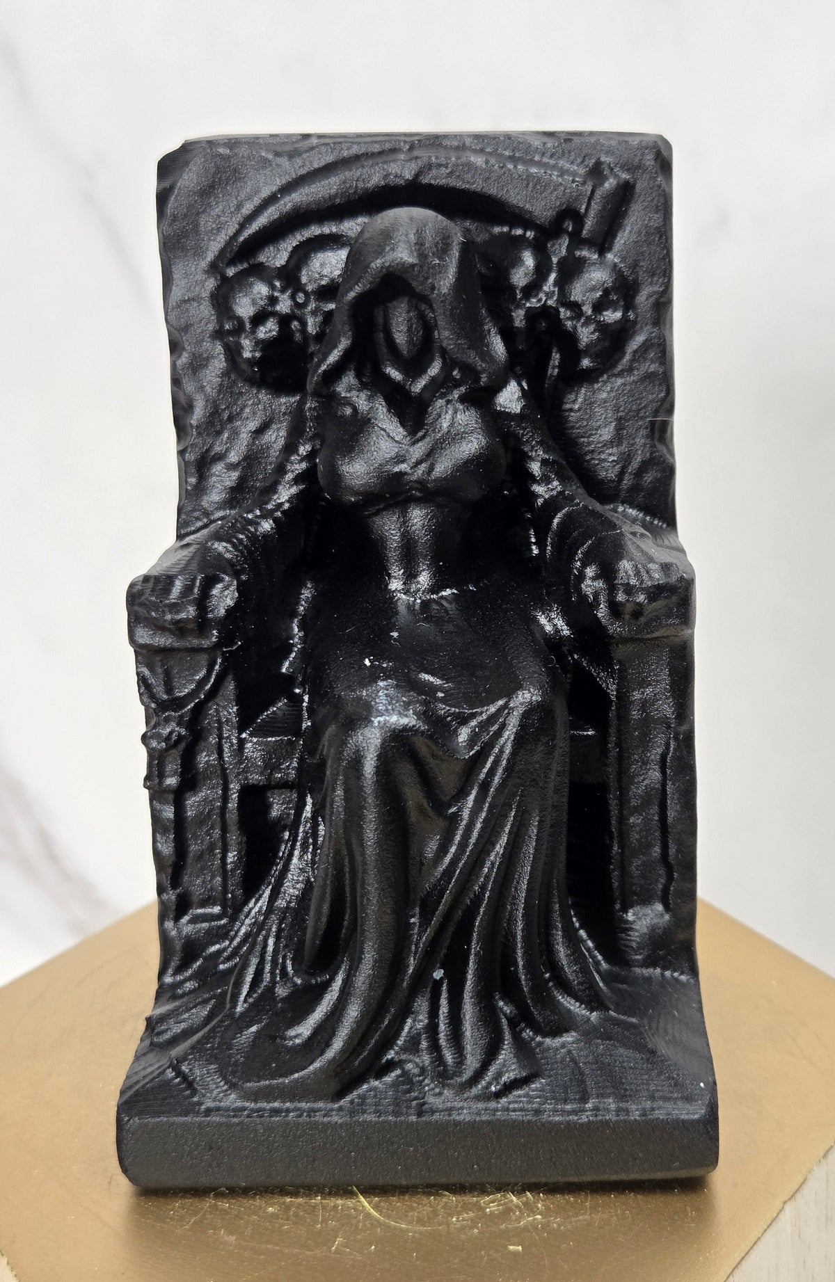 Carving of a faceless woman wearing a robe, sitting on a chair, in black obsidian, on a wooden stand against a marble background