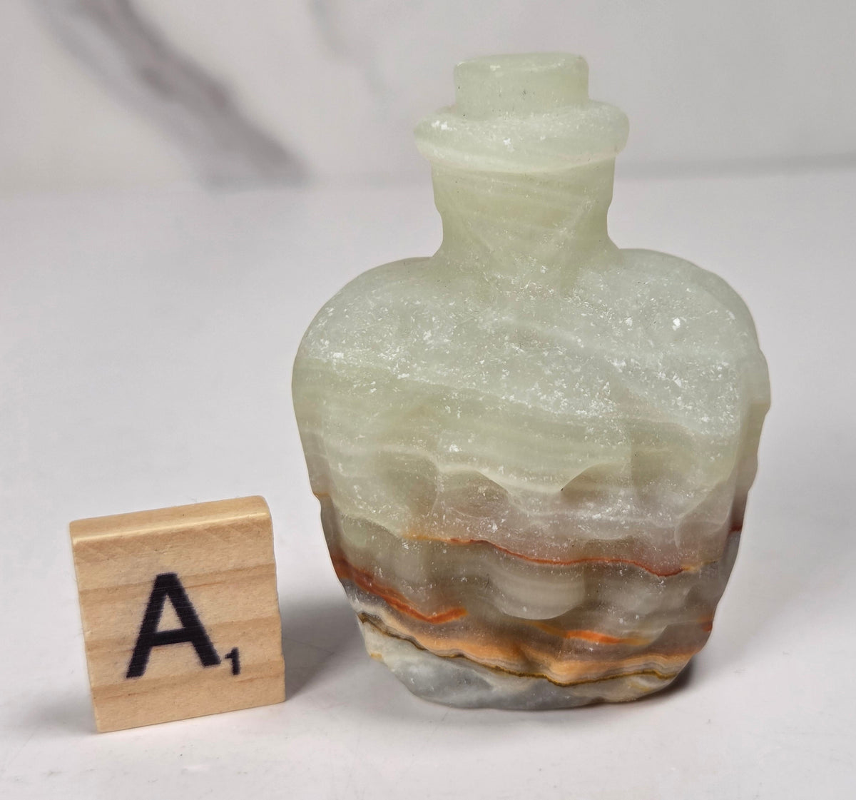 A small crystal bottle shaped like a skull, with a narrow neck and stopper, photographed on a plain light background with a scrabble wood tile.