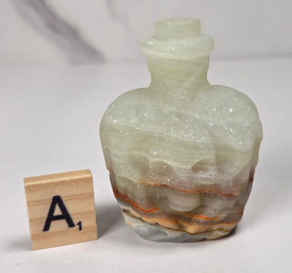 A small crystal bottle shaped like a skull, with a narrow neck and stopper, photographed on a plain light background with a scrabble wood tile.