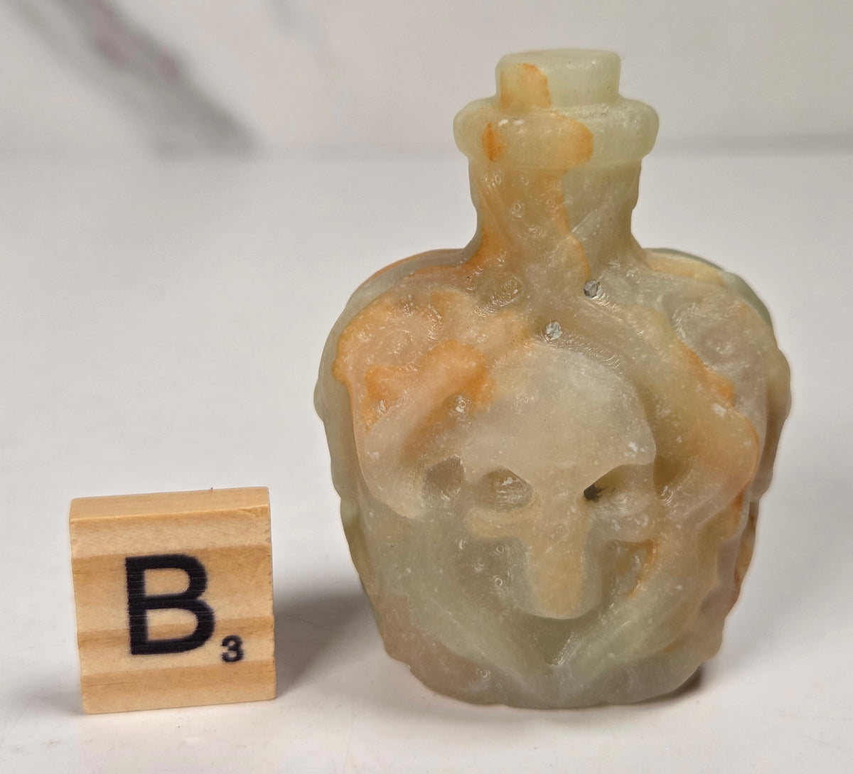 A small crystal bottle shaped like a skull, with a narrow neck and stopper, photographed on a plain light background with a scrabble wood tile.