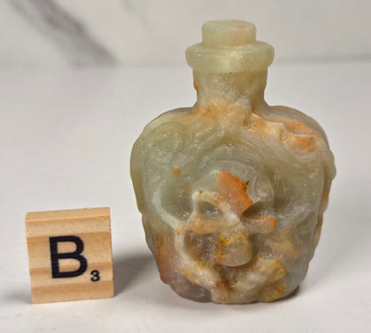 A small carved stone bottle with a skull motif, shown next to a wooden Scrabble tile displaying the letter B3 on a light marble-like surface.