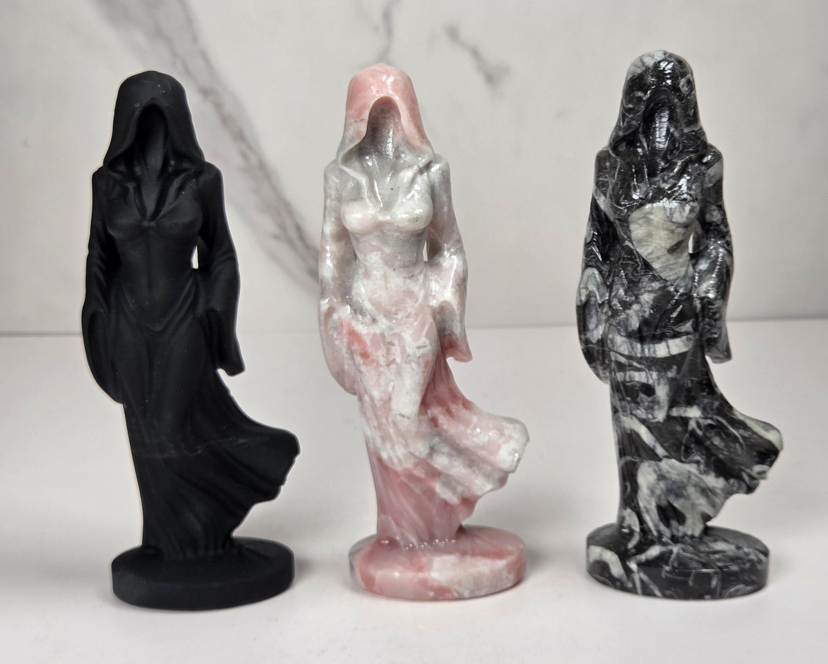 Set of 3 crystal carvings of a faceless woman wearing a flowy robe in varying colors of black, white and pink against a white background