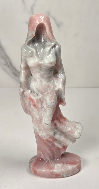 crystal carving of a faceless woman wearing a flowy robe with pink, white, and grey coloring against a white background