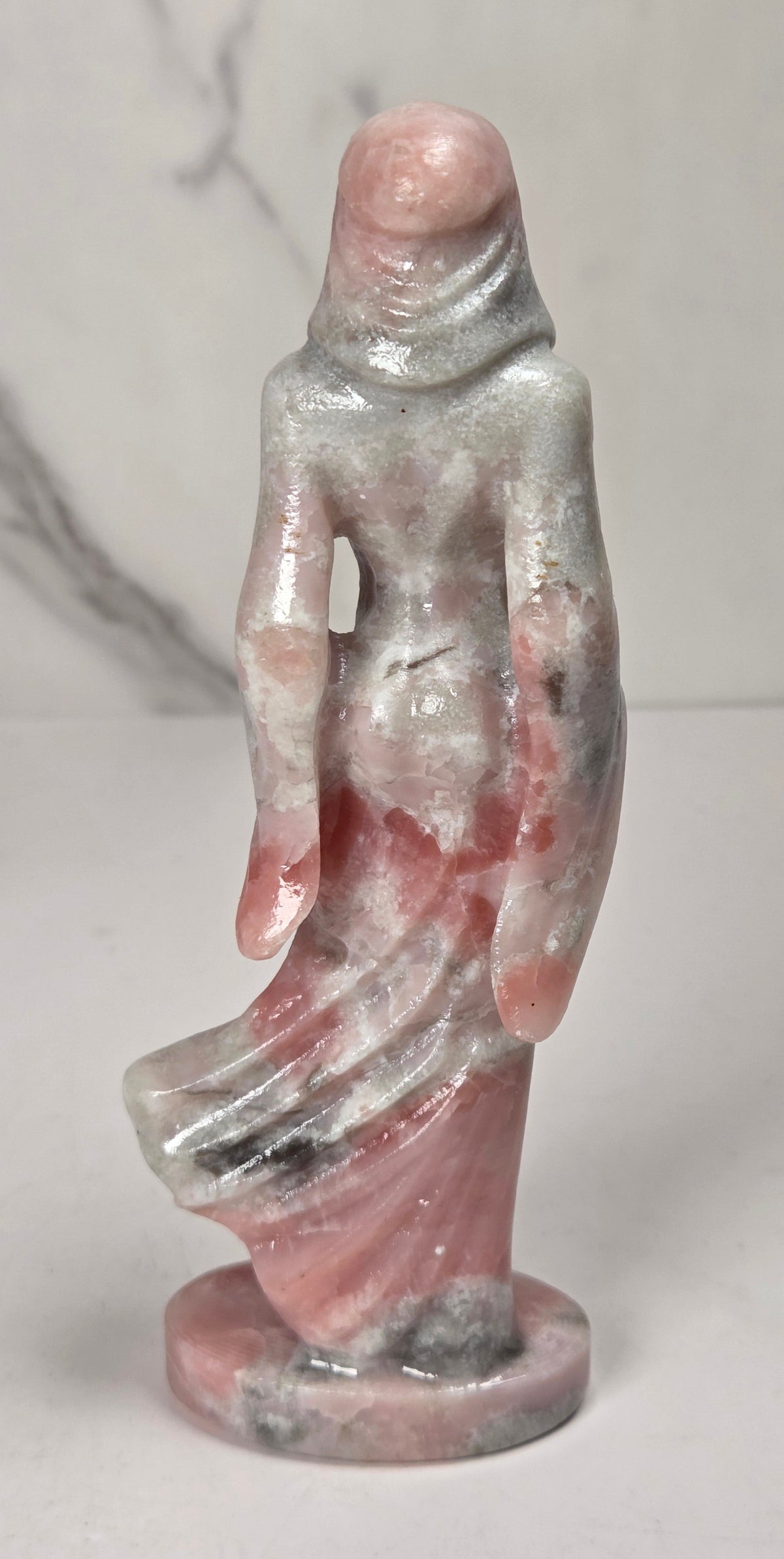 crystal carving of a faceless woman wearing a flowy robe with pink, white, and grey coloring against a white background
