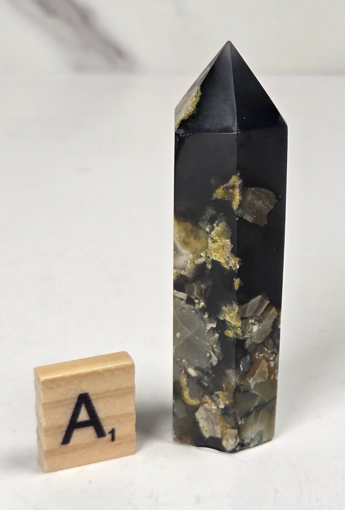 Volcano Agate polished point with tan and black coloring with a scrabble letter against a marbled background