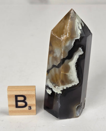 Volcano Agate polished point with tan and black coloring with a scrabble letter against a marbled background