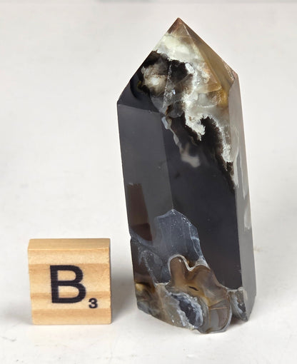 Volcano Agate polished point with tan and black coloring with a scrabble letter against a marbled background