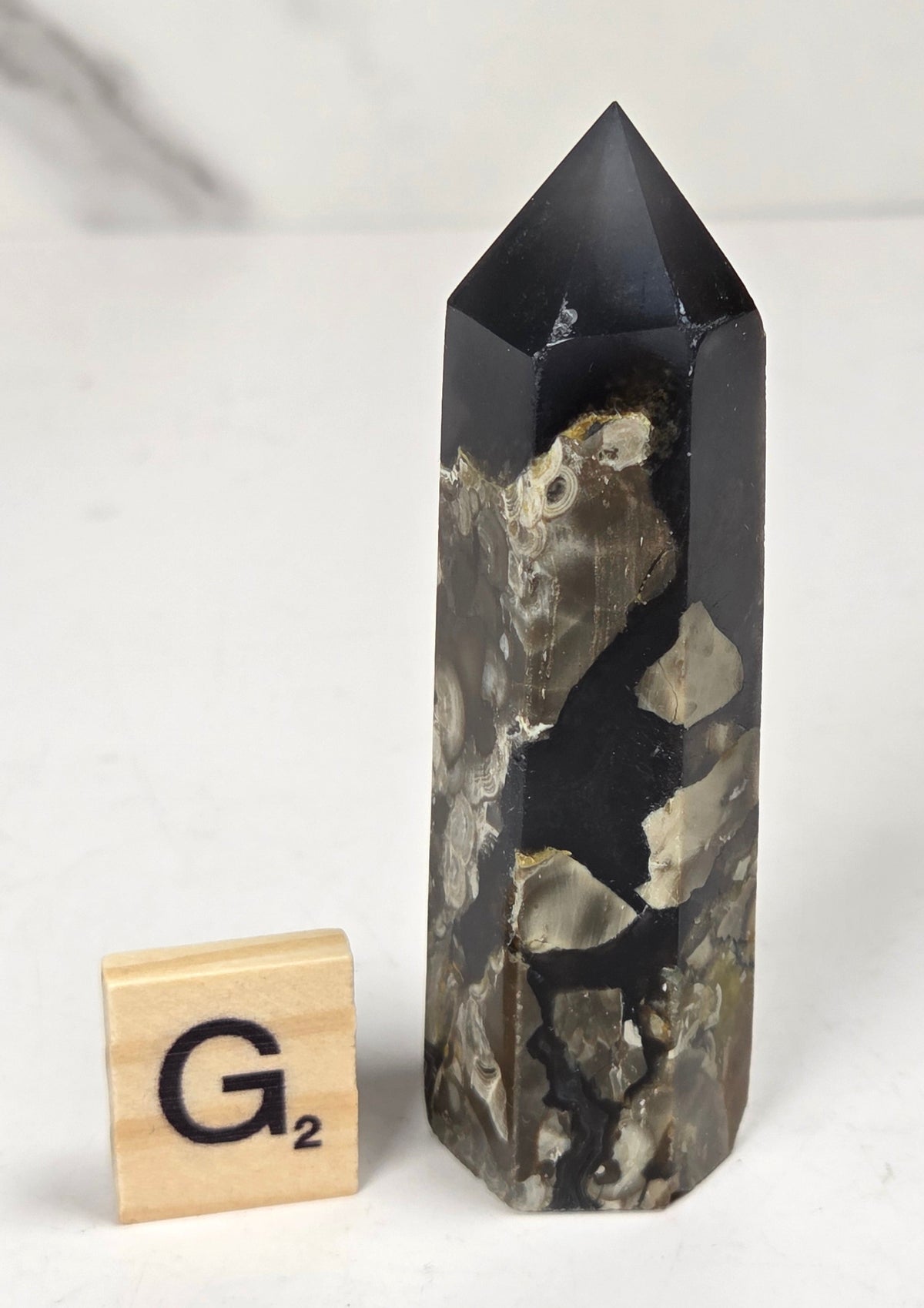 Volcano Agate polished point with tan and black coloring with a scrabble letter against a marbled background