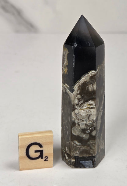 Volcano Agate polished point with tan and black coloring with a scrabble letter against a marbled background